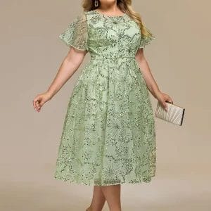 A woman wearing a light green, floral lace midi dress, holding a beige clutch, and standing in heeled sandals against a plain background.
