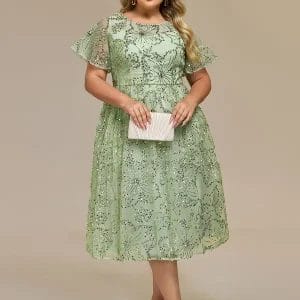 Woman wearing a light green knee-length lace dress with short flutter sleeves, holding a light-colored clutch, and wearing strappy heels, standing against a plain background.