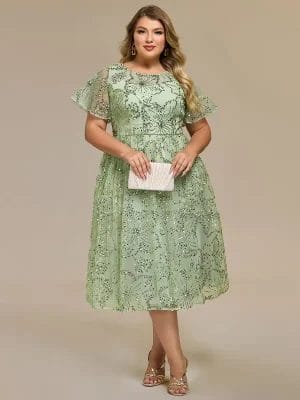 Woman wearing a light green knee-length lace dress with short flutter sleeves, holding a light-colored clutch, and wearing strappy heels, standing against a plain background.